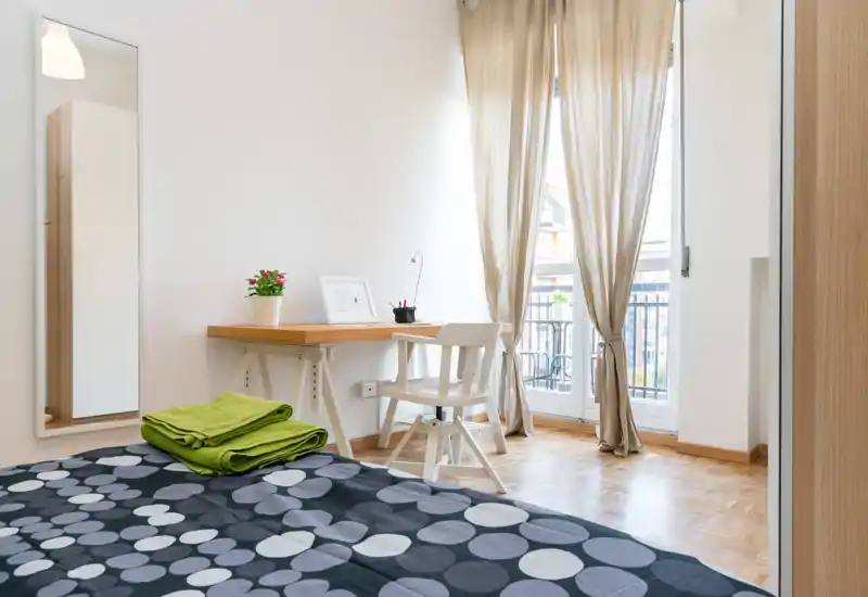 Dormitorio luminoso y cuidado con ropa de cama estampada, un pequeño escritorio de madera y silla junto a un balcón soleado. Tonos neutros, cortinas translúcidas y una planta decorativa transmiten una atmósfera tranquila y acogedora.