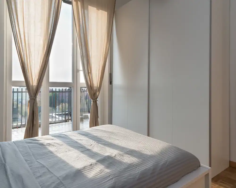 Dormitorio minimalista y luminoso con una cama doble bien hecha, ventanas de suelo a techo y cortinas neutras que dejan entrar luz natural cálida.