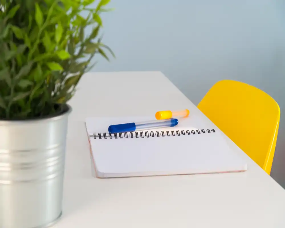 Primer plano de un rincón de escritorio ordenado con un cuaderno en espiral abierto, dos bolígrafos y una silla amarilla; una planta en maceta añade un toque de verde.