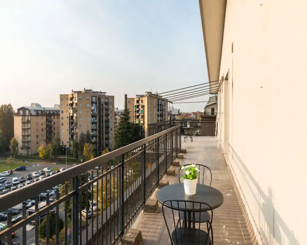 Amplio balcón soleado con barandilla metálica, pequeña mesa de bistró y sillas — ideal para el café de la mañana con vistas a los edificios vecinos y la calle arbolada.