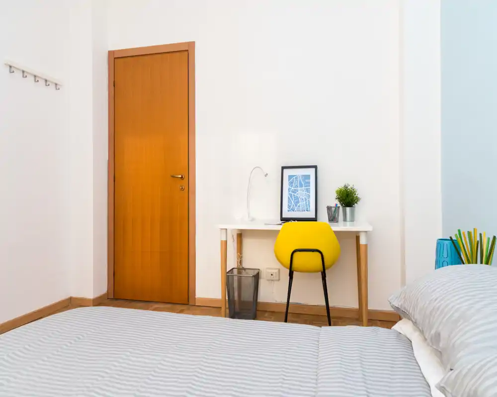 Dormitorio luminoso y sencillo con cama a rayas en primer plano, un pequeño escritorio blanco y una moderna silla amarilla contra una pared blanca. Perfecto para estancias individuales o cortas gracias al espacio de trabajo ordenado y la luz natural.