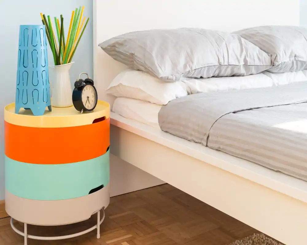 Rincón de dormitorio moderno y luminoso con cama bien hecha, ropa de cama gris a rayas y una mesita apilable de colores con accesorios decorativos.