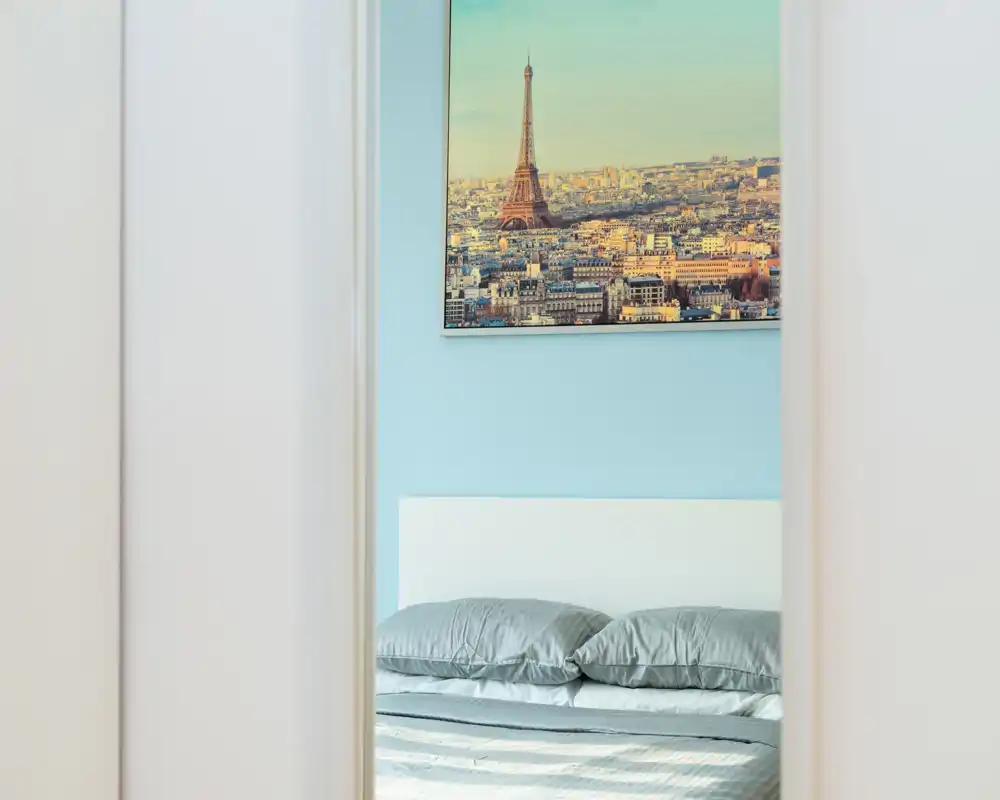 Dormitorio luminoso y minimalista visto a través de una puerta, con una cama doble bien hecha, almohadas grises y un cuadro de París sobre la cabecera.