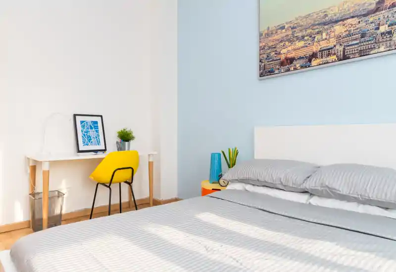 Dormitorio moderno y luminoso con una cama doble bien hecha, ropa de cama a rayas y un pequeño escritorio con una silla amarilla junto a la pared. La habitación recibe buena luz natural, decoración sencilla y un cuadro encima de la cabecera.