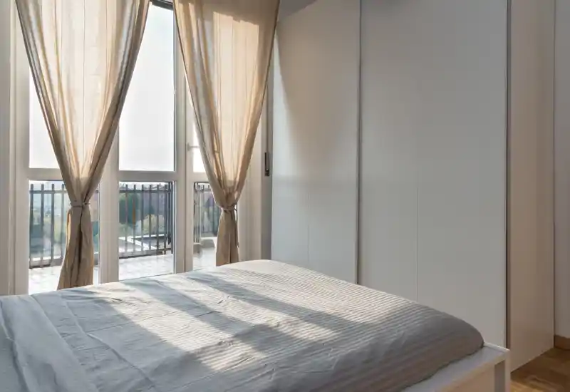 Dormitorio minimalista y luminoso con una cama doble bien hecha, ventanas de suelo a techo y cortinas neutras que dejan entrar luz natural cálida.