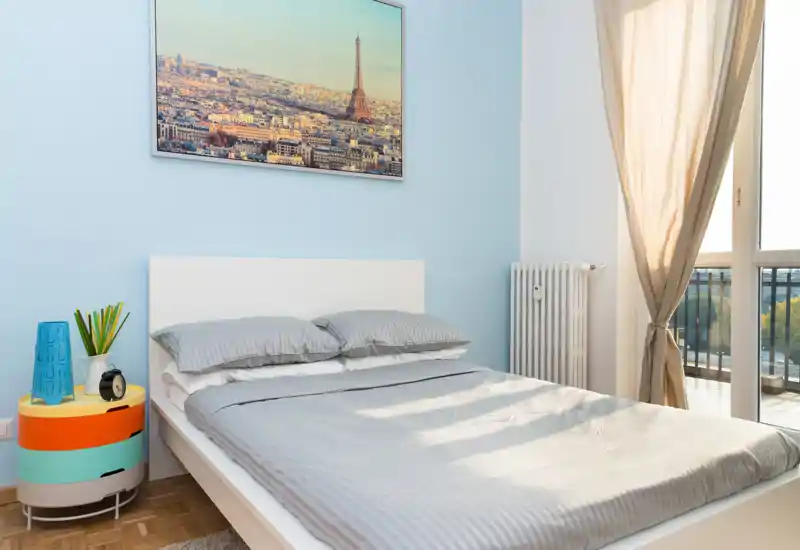 Dormitorio moderno y luminoso con cama doble vestida con ropa de cama gris, mesita de noche colorida y gran ventana que deja entrar luz natural. Paredes azul claro y cuadro de París sobre la cabecera.