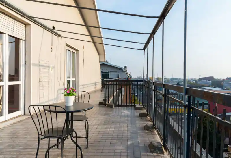 Grande terrasse sur le toit avec garde-corps métallique, sol carrelé et petite table avec deux chaises — idéale pour un café matinal et la vue sur la ville.