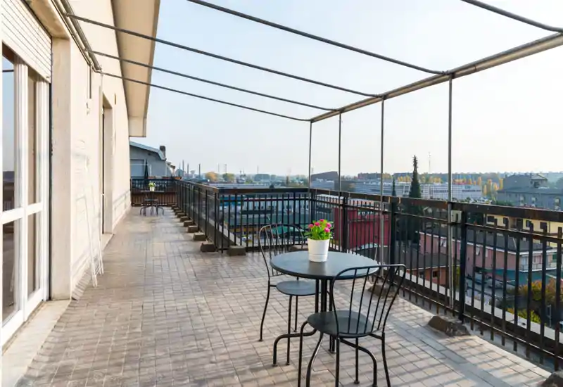 Grande terrasse sur le toit avec sol carrelé, garde-corps en métal et petite table avec chaises donnant sur la ville — espace extérieur lumineux, parfait pour se détendre ou déjeuner en plein air.
