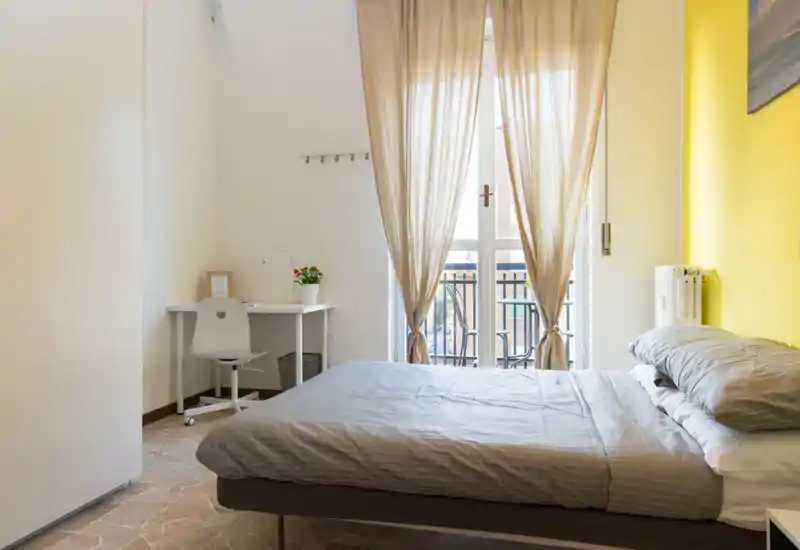 Bright, neatly staged bedroom with a comfortable double bed, soft grey linens, a small desk by the window and warm yellow accent wall letting in abundant natural light.