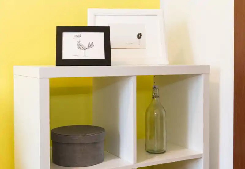 Bright, minimal corner of a living area with a white cube shelf, decorative framed prints, a fabric storage box and a glass bottle against a yellow accent wall.