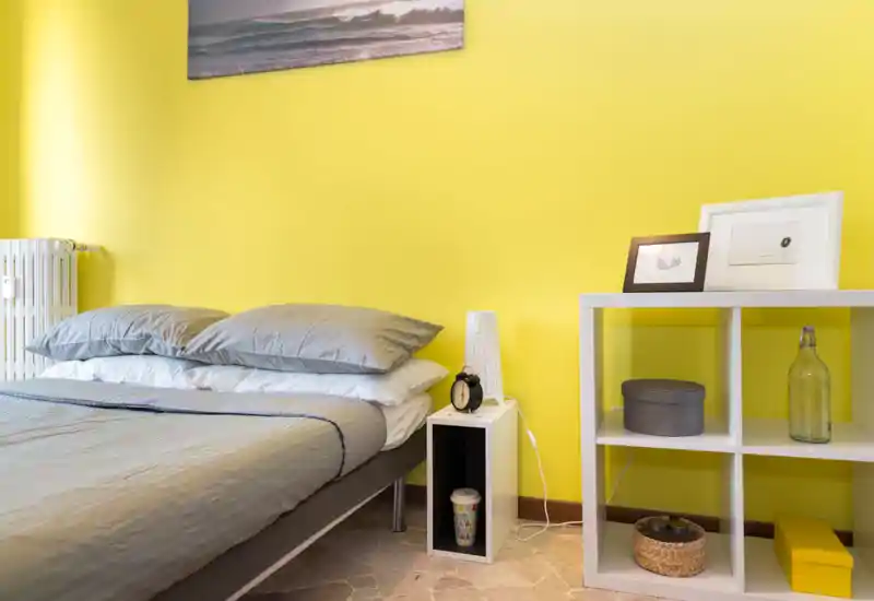 Sunny, modern bedroom with a neatly made double bed, soft grey linens and bright yellow walls; bedside shelf and decorative storage add practical styling for short-term rentals.
