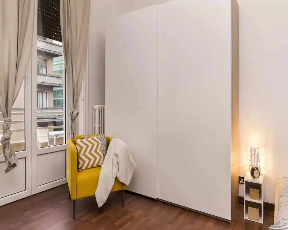 Stylish bedroom corner with a large white wardrobe, a bright yellow accent armchair and soft textiles; a small bedside table with a warm lamp and a balcony door providing daylight.