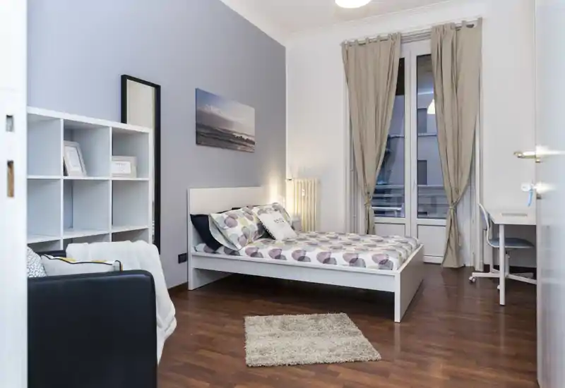 Dormitorio doble luminoso y bien decorado con cama blanca, ropa de cama estampada, pequeña alfombra y escritorio junto a la ventana — ideal para alquileres temporales o estudiantes.