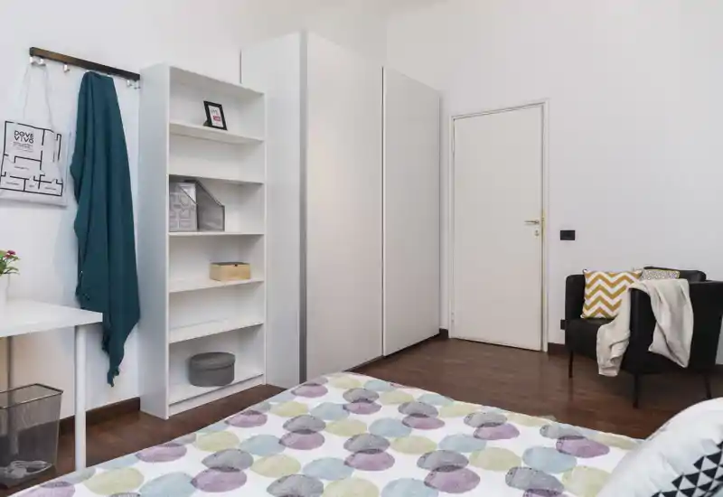 Dormitorio moderno y luminoso con colcha estampada, armario y estantería blancas, y un sillón acogedor junto a la puerta.