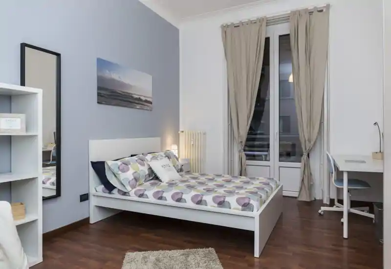 Dormitorio luminoso y bien decorado con cama doble, ropa de cama estampada, lámpara de noche y zona de trabajo junto a la ventana. Suelo de madera y cortinas neutras crean un ambiente tranquilo y acogedor perfecto para alquileres.