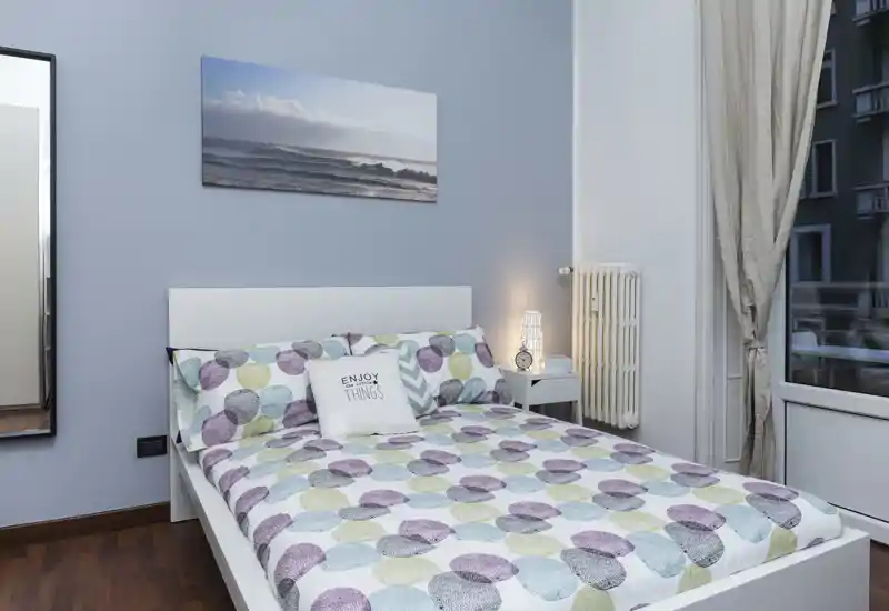 Dormitorio luminoso y bien decorado con cama doble y ropa de cama estampada, mesita con lámpara y una gran ventana que aporta luz natural — moderno y acogedor.