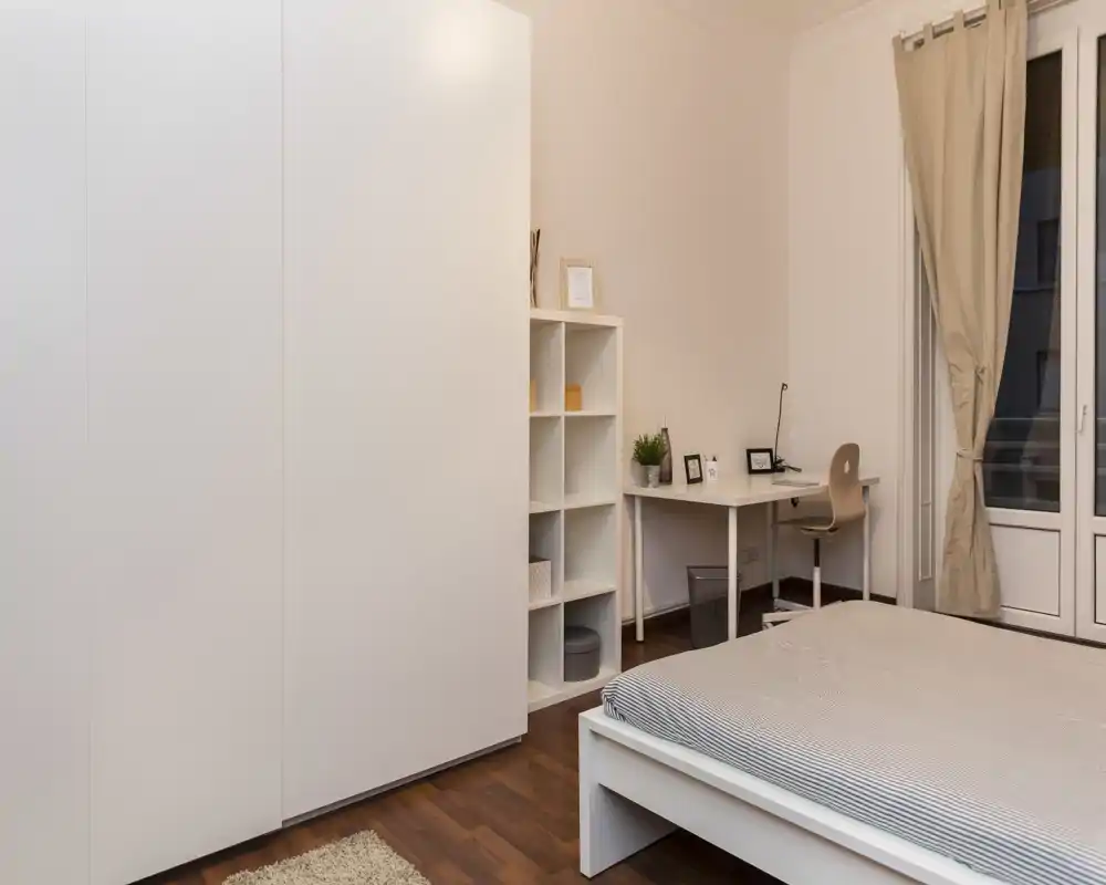 Dormitorio limpio y minimalista con estructura de cama blanca, amplio armario y un pequeño escritorio junto a la ventana; tonos neutros y suelo de madera cálido.