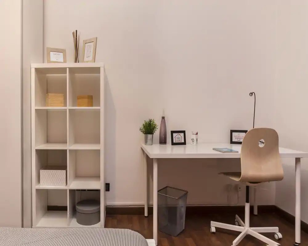 Rincón de dormitorio luminoso y minimalista con un escritorio sencillo y estantería. Se ve el borde de la cama en primer plano; la zona de trabajo incluye una planta, marcos y un escritorio blanco limpio.