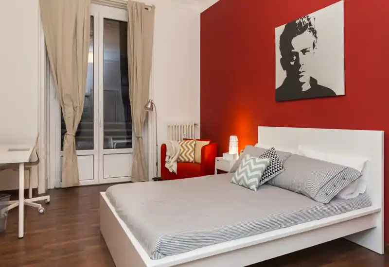 Dormitorio luminoso y con estilo: cama blanca, cojines estampados, lámpara de mesita y pared roja de acento que crean un ambiente moderno y acogedor.