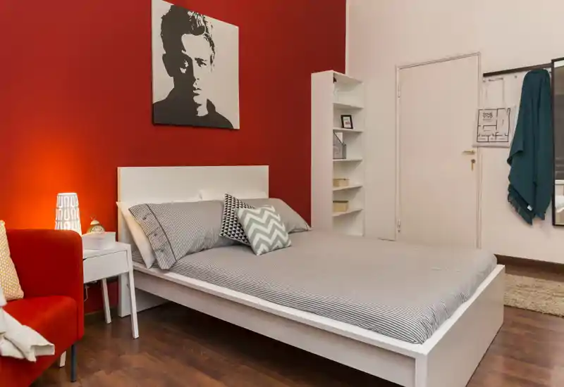 Dormitorio con estilo, cama de estructura blanca, ropa de cama con estampados y una llamativa pared roja. La habitación está cálidamente iluminada y bien decorada con cojines, una lámpara de noche y un sillón acogedor.