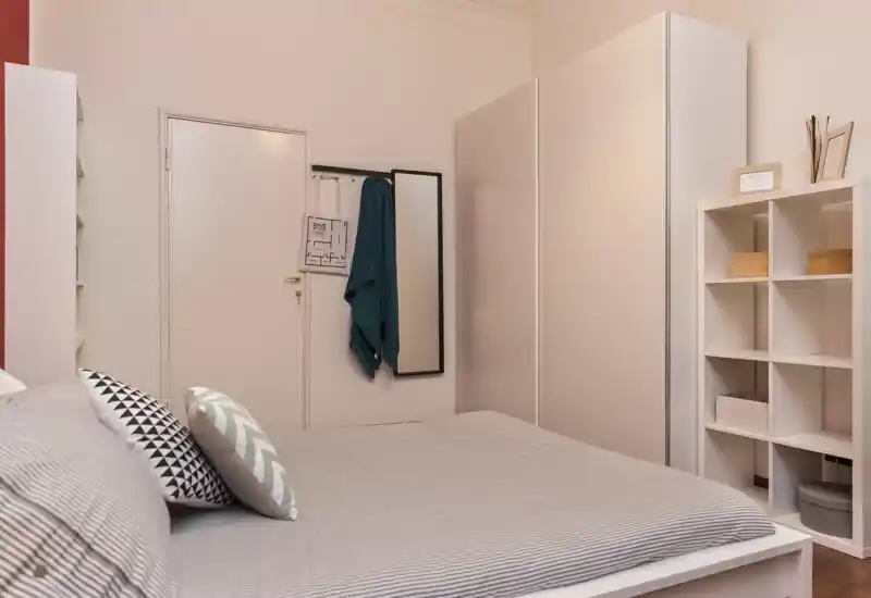 Dormitorio cómodo y bien decorado con una cama grande en primer plano, cojines decorativos, armario y estantería en la pared — tonos cálidos y espacio ordenado.