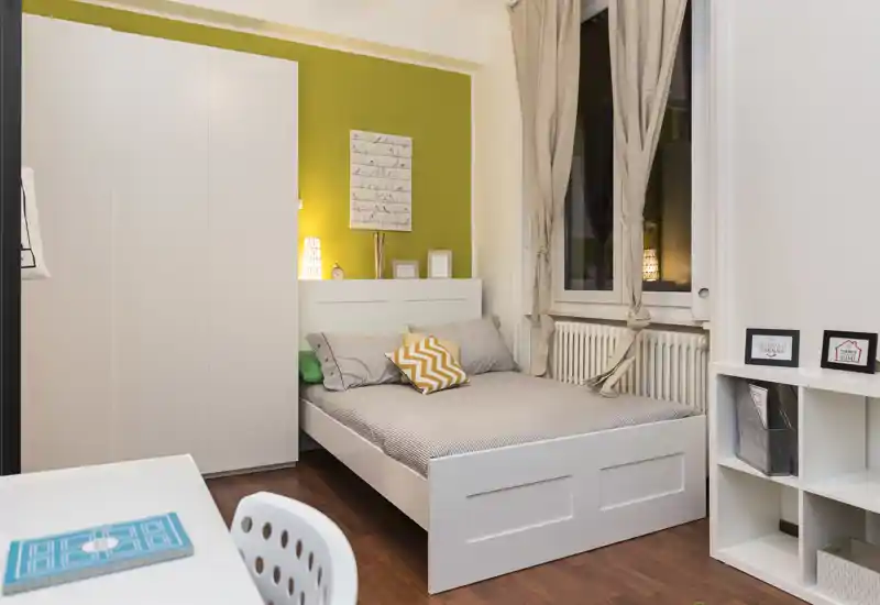 Dormitorio luminoso y bien decorado con cama blanca, textiles cálidos y pared verde de acento — ideal para fotos de alquiler que muestren una zona de descanso acogedora.