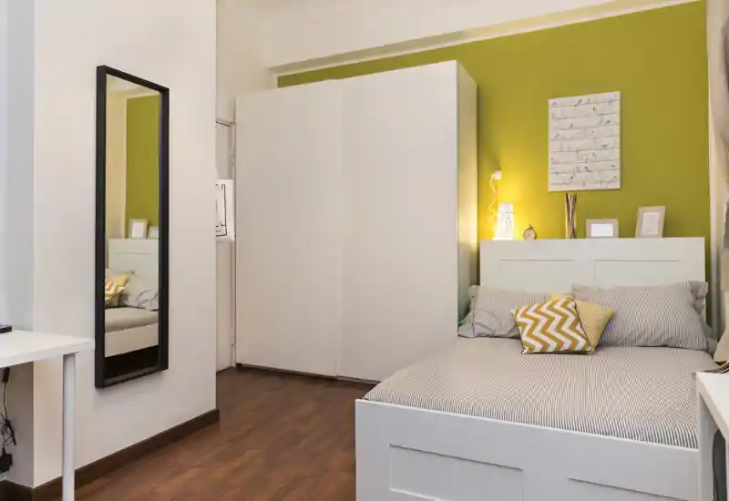 Dormitorio luminoso y bien decorado con cama blanca, cojines decorativos y un gran armario frente a una pared verde — ambiente moderno y acogedor.