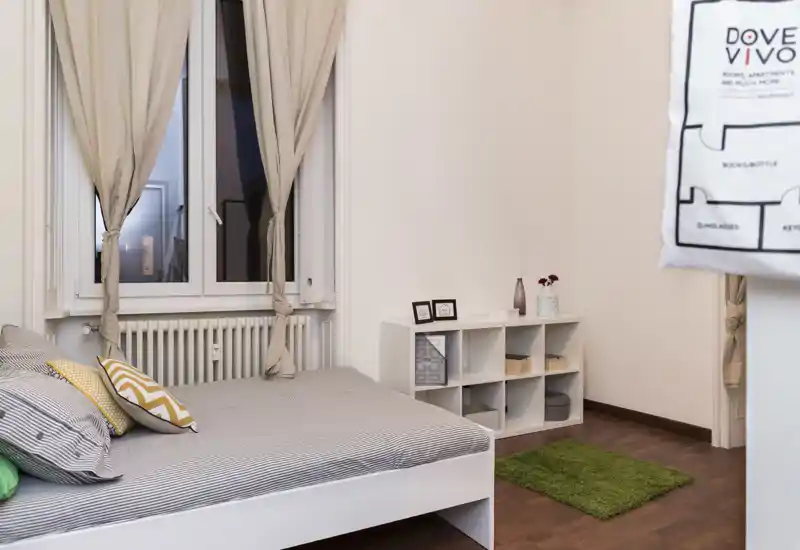 Dormitorio ordenado y bien iluminado con cama doble vestida con ropa de cama a rayas, cojines decorativos y cortinas claras en la ventana. Estantería baja y una alfombra verde aportan almacenamiento y un toque de color.