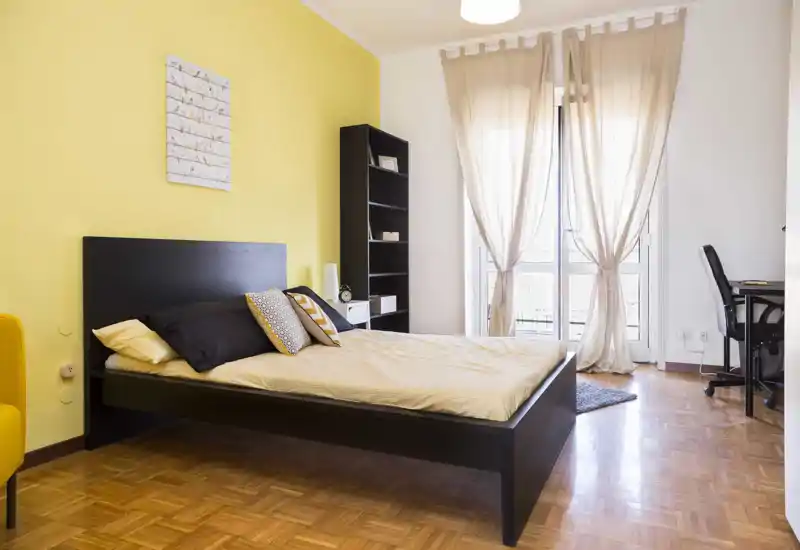 Dormitorio moderno y luminoso con una cama de estructura negra, pared amarilla de acento y cortinas ligeras que dejan pasar la luz natural — habitación acogedora y bien presentada.
