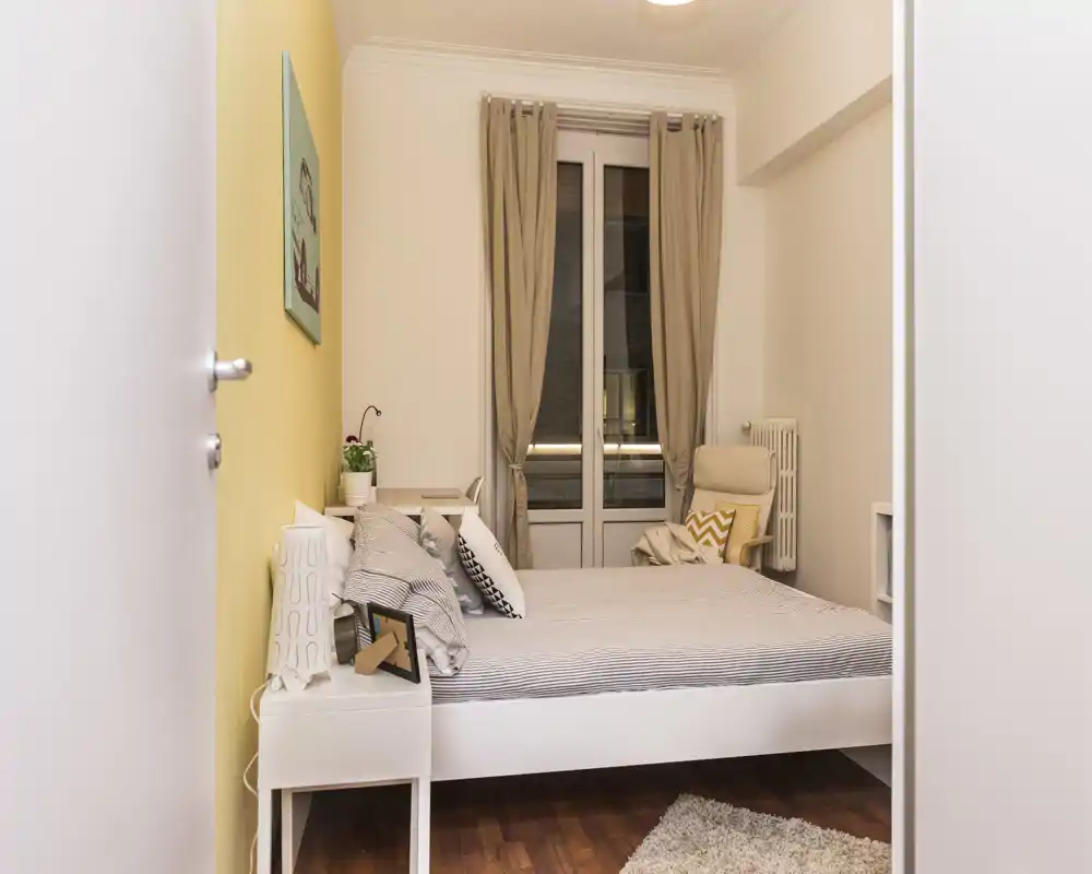 Dormitorio luminoso y ordenado con cama doble, textiles suaves y un cómodo sillón junto a la ventana — ideal para anuncios de alquiler.