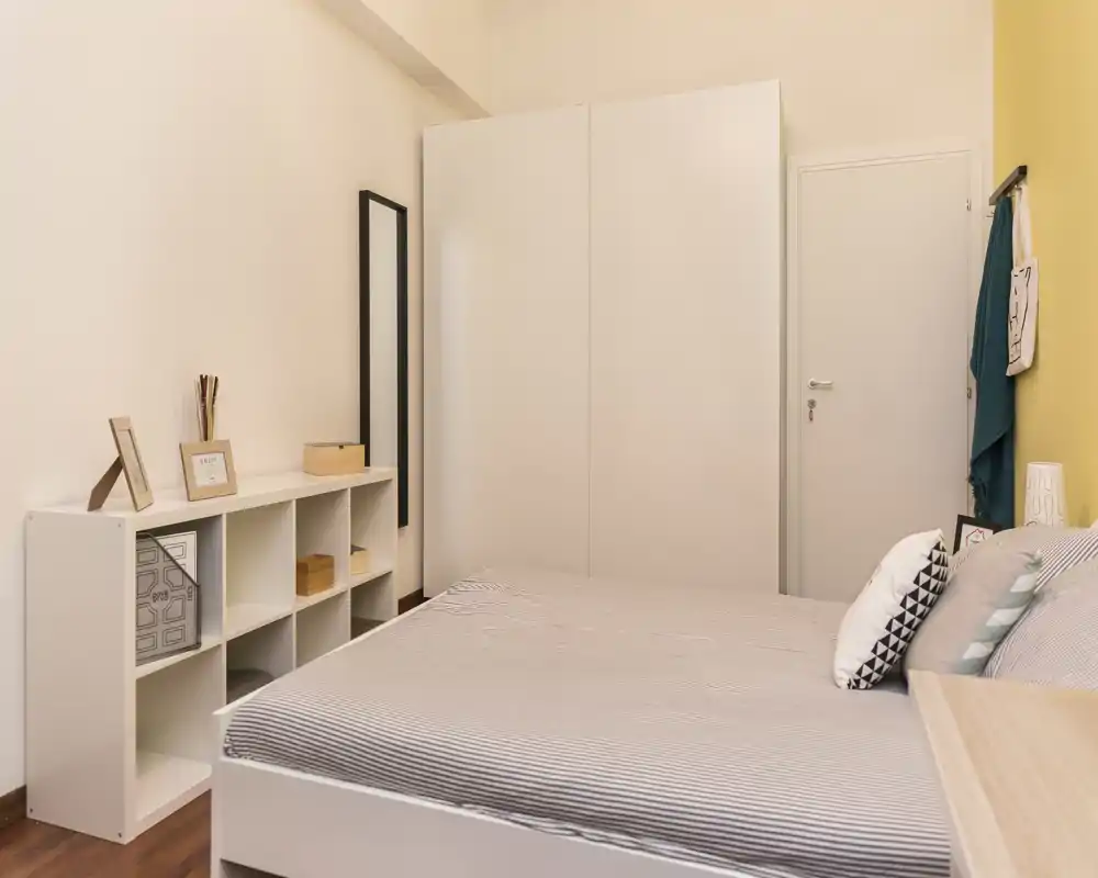 Dormitorio luminoso y bien decorado con cama doble en primer plano, armario blanco y estantería; tonos neutros que transmiten calma.