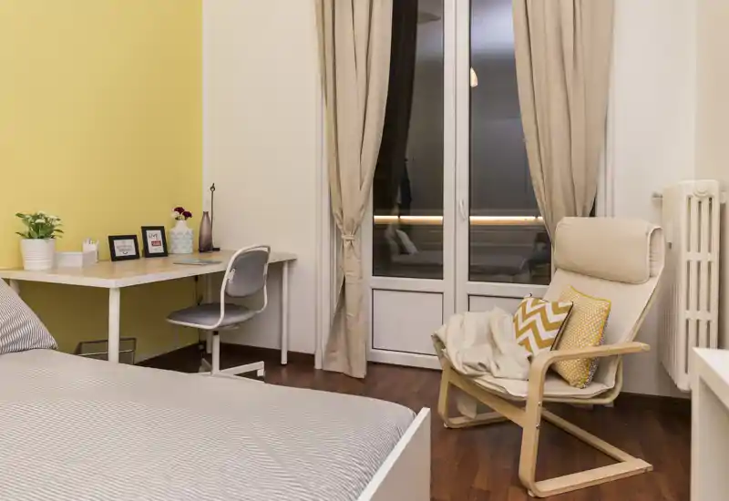 Dormitorio moderno y acogedor con la cama bien hecha, un escritorio junto a una pared amarilla y un sillón cómodo junto a la puerta-balconcillo.