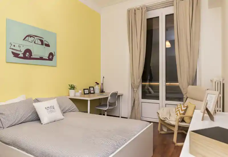 Dormitorio luminoso y ordenado con cama doble, cojines decorativos y un pequeño escritorio junto a la ventana. Tonos neutros y pared amarilla que aportan un ambiente acogedor y moderno.