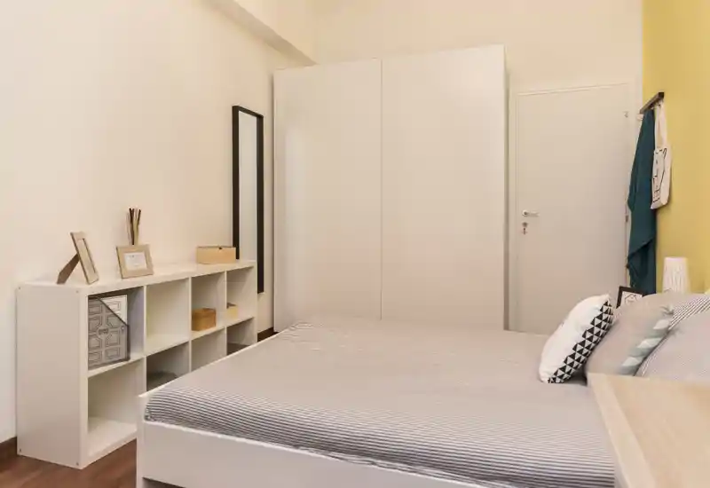 Dormitorio luminoso y bien decorado con cama doble en primer plano, armario blanco y estantería; tonos neutros que transmiten calma.