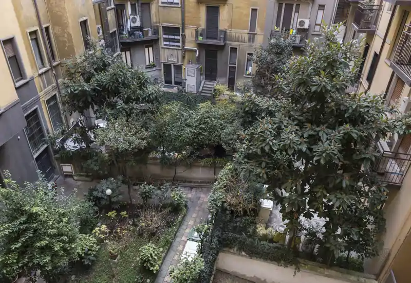 Inner courtyard garden with mature trees and planted beds, viewed from an upper floor. The space offers greenery and a paved path between residential buildings.