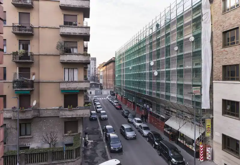 Street-facing view from a window showing parked cars, apartment facades and a building under renovation with scaffolding — ideal to illustrate the neighbourhood and immediate surroundings.