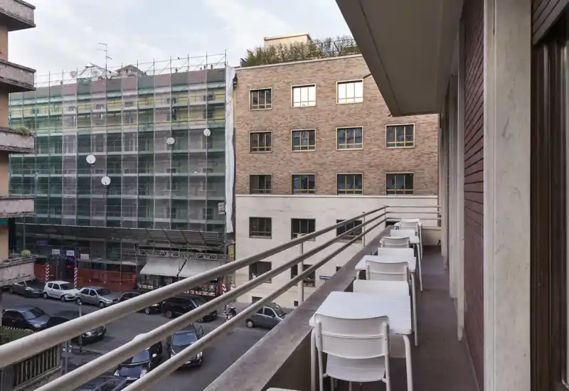 Sunny urban balcony with small white tables and chairs, overlooking a street and neighboring buildings — ideal for morning coffee or light outdoor seating.