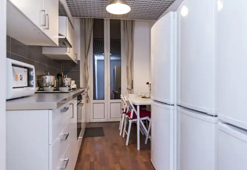 Cocina moderna y estrecha con muebles blancos, electrodomésticos y un pequeño rincón comedor con dos sillas — luminosa y ordenada, perfecta para cocinar de forma eficiente.