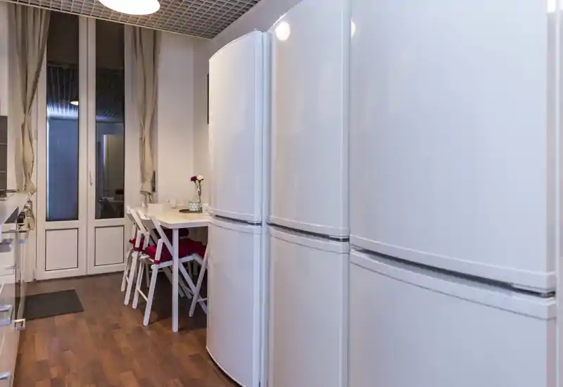 Contemporary galley kitchen with white appliances, a compact dining nook with chairs and cushions, and wood flooring — bright and neatly arranged for everyday use.
