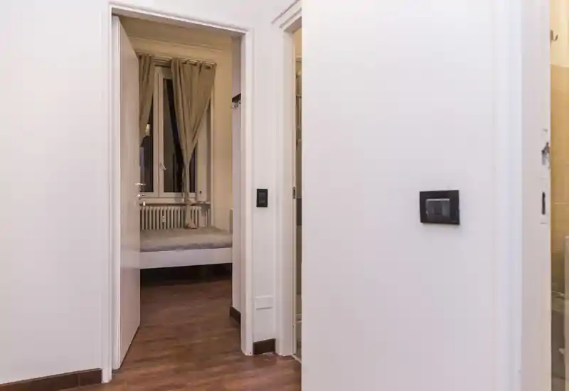 Vista desde el pasillo hacia un dormitorio compacto con cama visible, suelos de madera y cortinas neutras; luminoso, limpio y de mobiliario sencillo, ideal para alquileres cortos o habitación de invitados.
