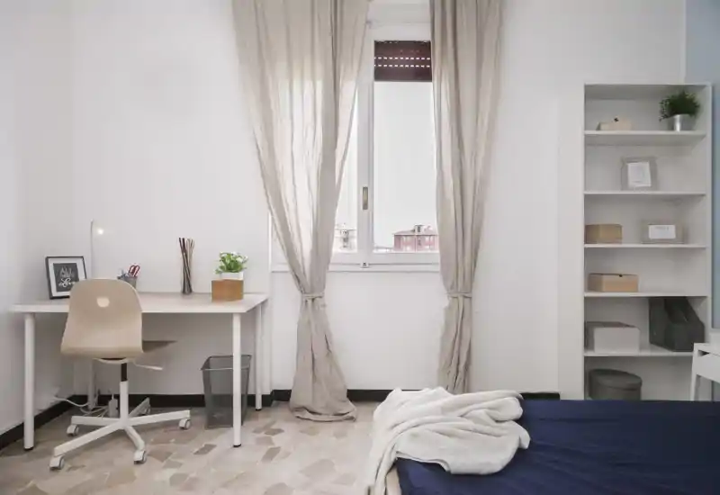 Bright, minimalist bedroom with a visible bed in the foreground, a desk and chair by the window, and open shelving—ideal for a peaceful, work-friendly stay.