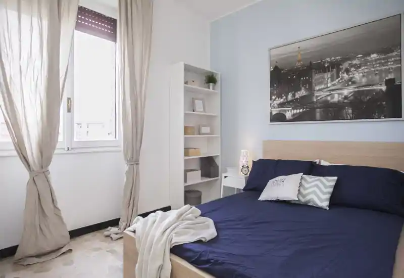 Bright, well-staged bedroom with a double bed dressed in deep navy linens, decorative cushions and a cozy throw. Natural light from a large window and tasteful wall art create an inviting, modern atmosphere.