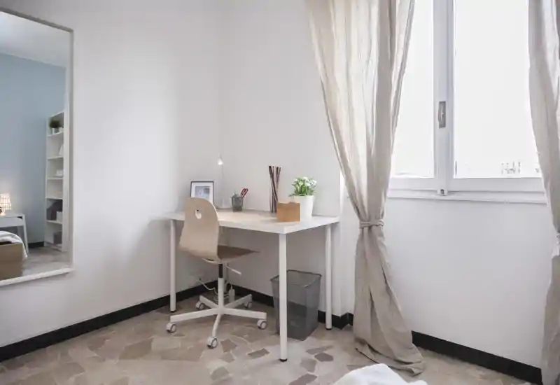 Bright, minimalist bedroom corner featuring a simple desk and chair by a large window; a bed is visible only as a reflection in the mirror, suggesting a compact sleeping area.