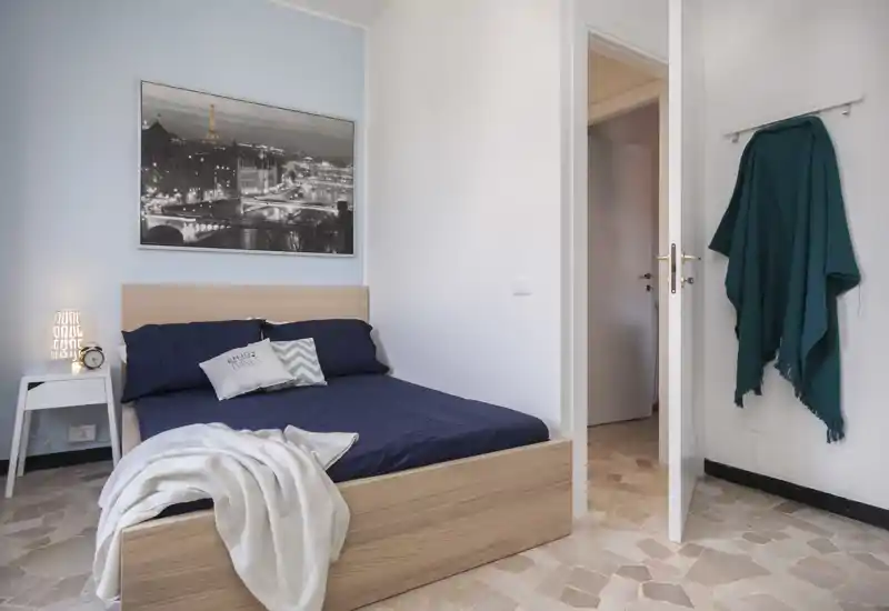 Bright, modern bedroom with a wooden frame bed dressed in navy linens, decorative pillows and a soft throw. Minimal nightstand with lamp and a monochrome city print above the headboard create a calm, cozy atmosphere.