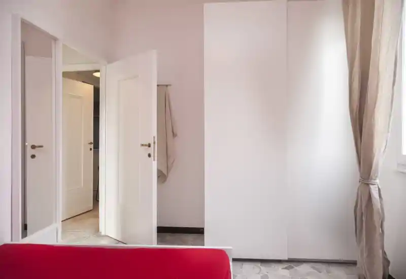 Rincón de dormitorio luminoso y minimalista con una cama cubierta de rojo en primer plano, un armario blanco y una ventana con cortinas que dejan entrar luz natural suave.