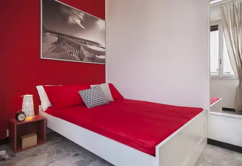Dormitorio moderno y luminoso con estructura de cama blanca y ropa de cama roja intensa, cojines decorativos y lámpara de mesita — bien iluminado y atractivo para alquileres.