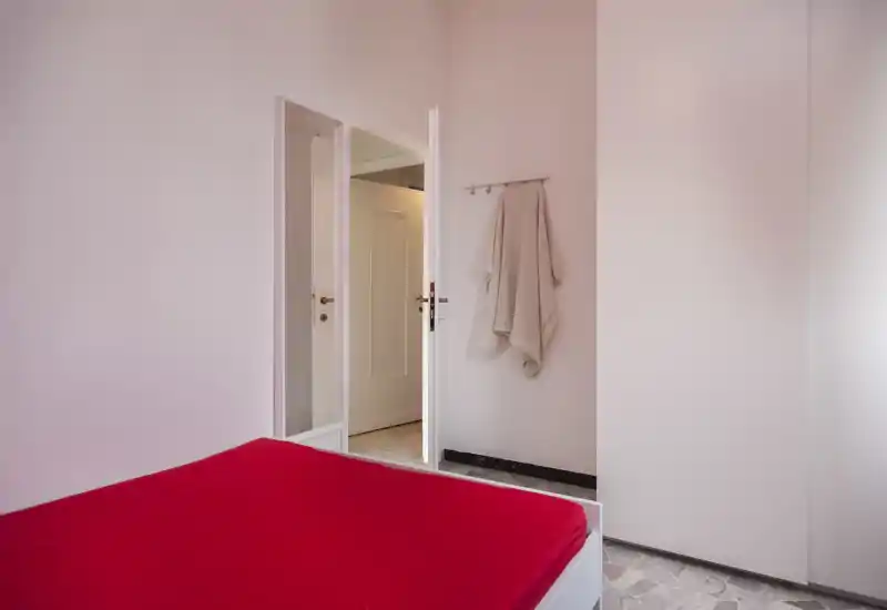 Dormitorio sencillo y luminoso con cama cubierta de rojo, paredes blancas y armario; ambiente limpio con luz natural y estilo minimalista.