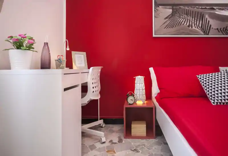 Rincón de dormitorio moderno y luminoso con cama en rojo, cojines geométricos, mesita con lámpara y despertador y un escritorio blanco compacto: un espacio acogedor y bien decorado para un huésped.