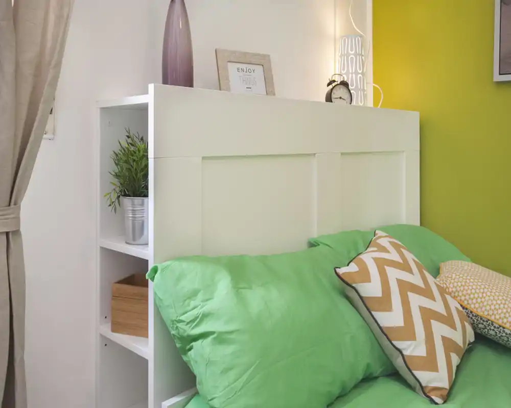 Rincón de dormitorio luminoso y cuidado con cabecero blanco, ropa de cama verde y cojines estampados; estantería decorativa con planta y marco que aporta calidez.