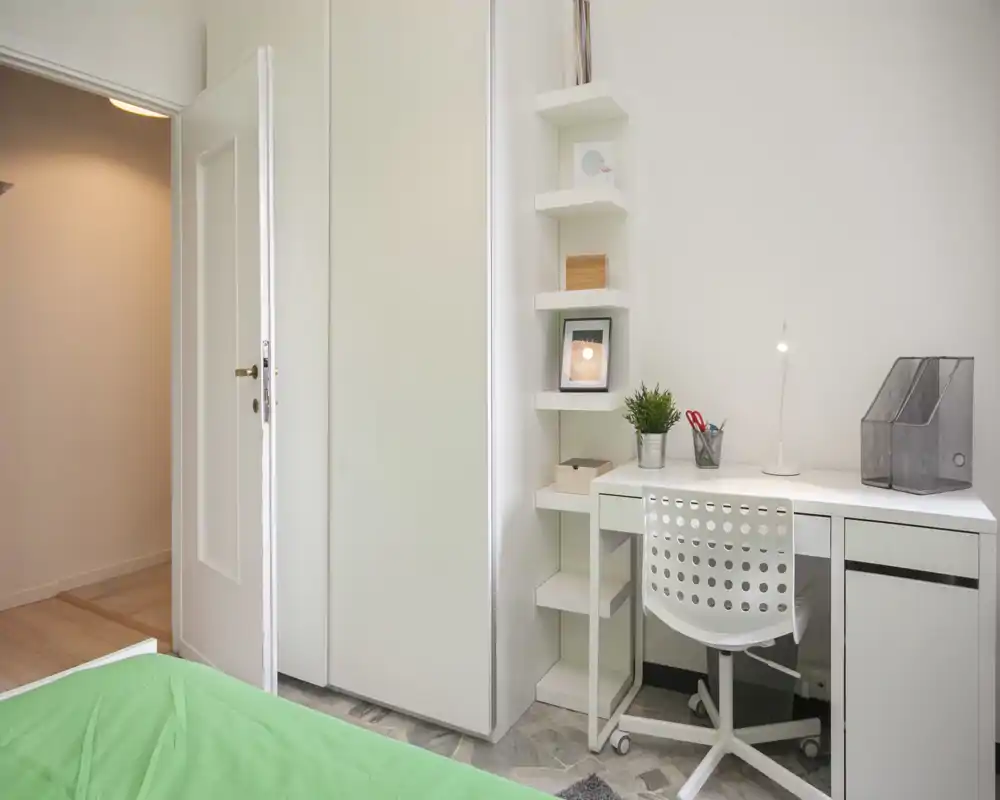 Rincón de dormitorio compacto con escritorio y silla blancos junto a un armario empotrado; se ve parcialmente una colcha verde en primer plano. Espacio luminoso, minimalista y ordenado, ideal para un estudiante o persona sola.